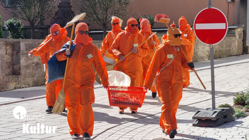 Vermummte und orange gekleidete Aktionskünstler mit Besen auf der Straße einer fränkischen Kleinstadt