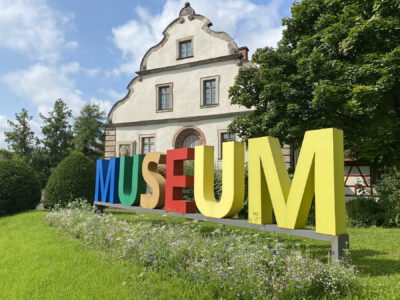 Bunter Schriftzug "Museum" im Garten vor dem historischen Museumsgebäude.