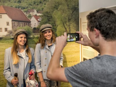 Eine Person fotografiert zwei junge Frauen, die ihre Köpfe durch eine Fotowand stecken und dadurch aussehen als wären sie in historischer Müllerkluft.