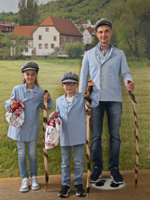Zwei Kinder und ein Erwachsener posieren in Müllerkluft mit Wanderstäben auf einer Wiese.