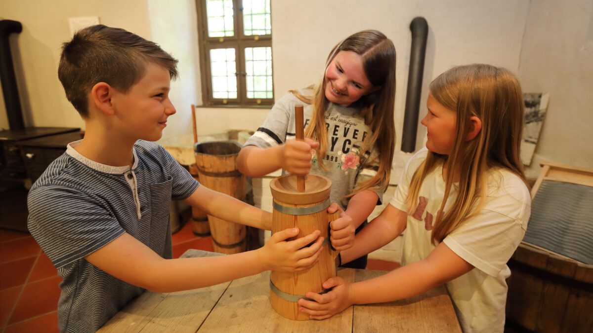 Drei Kinder stampfen Butter in einem historischen Butterfass.