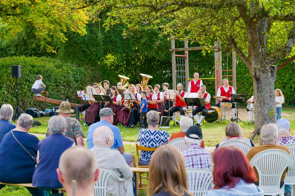 sitzende Zuhörer und spielende Kinder rund um eine Musikkapelle auf einem Spielplatz