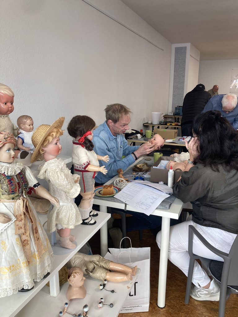 Im Vordergrund sind Puppen und Puppenersatzteile zu sehen. Im Hintergrund sitzt ein Mann mit einem Puppenkopf in der Hand.